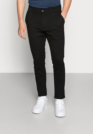 Man die slanke zwarte broek, marineblauw overhemd en witte sneakers draagt, staat met één hand in zijn zak tegen een effen achtergrond.