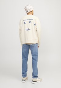 Sweatshirt crème avec du texte bleu dans le dos, coupe décontractée, associé à un jean bleu clair et des baskets beiges, porté avec un chapeau seau à motifs.