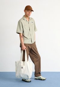 Camisa de manga corta verde claro, pantalones marrones, bolso de mano de rayas crema con logo, y zapatillas verde claro con suela negra.