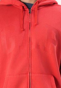 Crvena zip-up dukserica od mekog materijala s prednjim džepom, koja ima suptilan preveliki "GAR" logo u istoj boji.