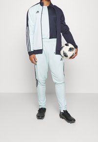 Hellblaues Adidas-Trainingsanzug mit schwarzen Akzenten, der drei Streifen auf den Ärmeln und der Hose aufweist. Schwarzer Fußball in der rechten Hand gehalten.