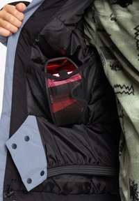 Schwarz-graues Innenfutter der Jacke mit einer Netztasche, die rote Sonnenbrillen hält. Der innere Stoff ist glatt; das äußere hat eine mit Knöpfen geschlossene Klappe und einen elastischen Bund.
