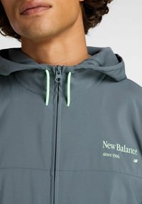 Hombre vestido con una chaqueta gris de New Balance, cerrada con cremallera, que presenta cordones de neón verde y el texto del logo "New Balance desde 1906" en el pecho.