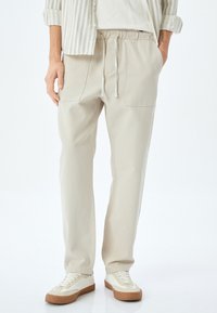 Beige, leichte Hose mit elastischem Bund und Kordelzug. Verfügt über zwei Fronttaschen und eine lockere Passform. Getragen mit weißen Sneakers.