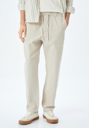 Pantalones ligeros color beige con cinturilla elástica y cordón ajustable. Presentan dos bolsillos frontales y un corte relajado. Llevados con zapatillas blancas.