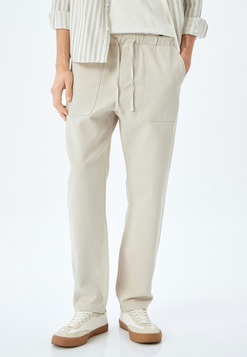 Beige, leichte Hose mit elastischem Bund und Kordelzug. Verfügt über zwei Fronttaschen und eine lockere Passform. Getragen mit weißen Sneakers.