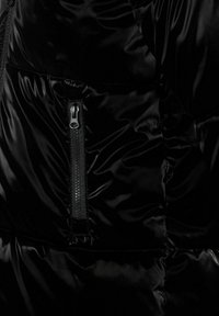 Secțiune a jachetei puffer negre, cu o textură netedă și lucioasă, având un buzunar cu fermoar și detalii de design quilted evidențiate.