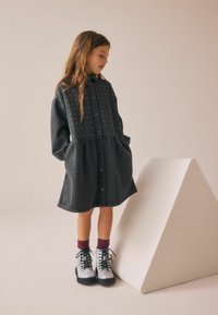 Abito camicia in denim nero con bottoni frontali, dettagli in perle sul corpino, vita arricciata e maniche lunghe. Abbinato a stivali argento scintillanti.