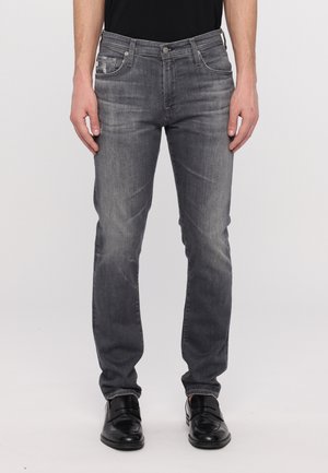 Mann trägt ausgewaschene graue Slim-Fit-Jeans und schwarze Anzugsschuhe, steht vor einem schlichten hellen Hintergrund.