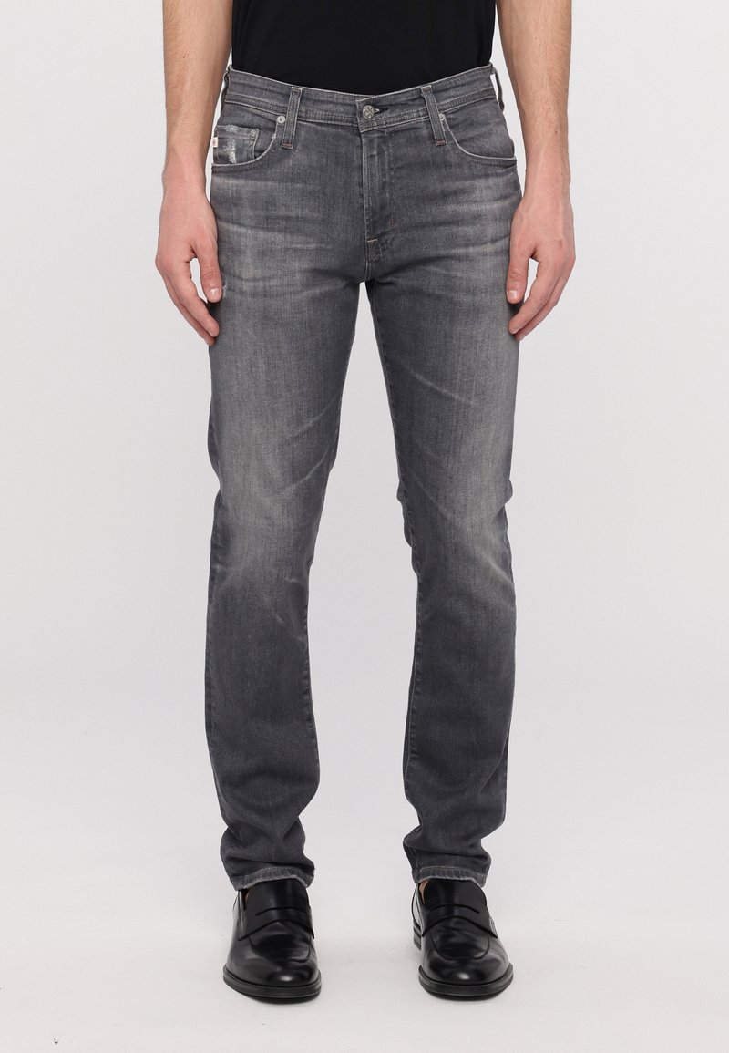 Homme portant un jean slim gris délavé et des chaussures habillées noires, debout devant un fond clair uni.