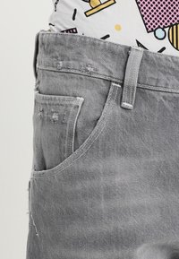 Primer plano de un bolsillo de jeans de mezclilla gris con un ligero desgaste y un pasador de cinturón, acompañado de una camisa de patrones coloridos metida por dentro.