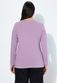 Langärmliges Oberteil in hellviolett, aus weichem Baumwollmaterial mit rundem Halsausschnitt und geradem Saum, kombiniert mit dunklen Hosen. Rückansicht dargestellt.