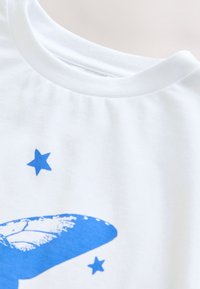 T-shirt en coton blanc avec un motif bleu représentant une baleine et deux étoiles bleues. Texture douce avec un col rond et des manches courtes.