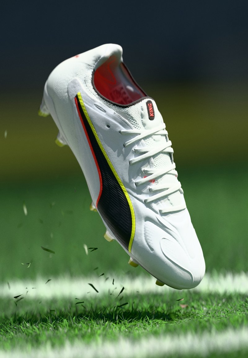 Scarpa da calcio bianca con dettagli neri e rossi, tomaia testurizzata, sei tacchetti gialli e marchio "KING" sulla linguetta. Ci sono fili d'erba visibili.