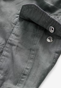 Grauer Stoff mit glatter Oberfläche, ausgestattet mit einem umgeschlagenen Kragen, sichtbaren Nähten und metallischen Druckknopfdetails. Zwei Taschen sind teilweise sichtbar.
