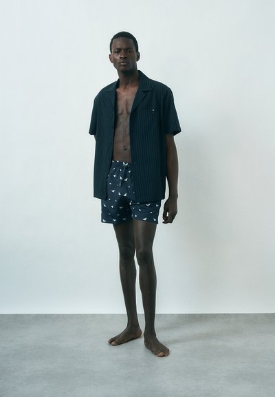 Άνδρας στέκεται ξυπόλητος, φοράει ανοιχτό σκούρο κοντομάνικο πουκάμισο και μπλε μαγιό με μοτίβα, πάνω σε απλό ανοιχτό φόντο.