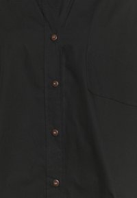 Camicia nera con bottoni marroni, taschino frontale e texture liscia. Il tessuto appare leggero e resistente.
