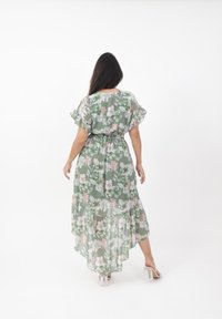 Abito maxi floreale in chiffon verde con orlo asimmetrico, maniche a volant e vita elastica. Presenta motivi floreali rosa e bianchi.