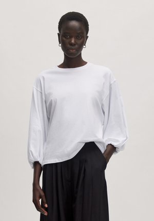 T-shirt blanc à manches longues avec un col rond et des manches volumineuses, associé à un pantalon noir plissé taille haute.