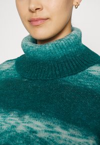 Turkizna pulover s turtleneck ovratnikom in rebrasto teksturo, ki ima gradientni vzorec in mehak, rahlo puhast material. Poudarjen je detajl na ovratniku.