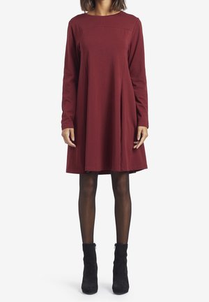 Abito lungo bordeaux a maniche lunghe con scollatura rotonda, silhouette morbida e orlo a ginocchio. Abbinato a stivali neri alla caviglia e collant scuri.