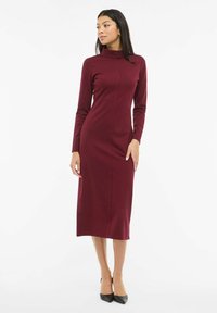 Vyno raudonio spalvos su ilgomis rankovėmis ir stovi kaklo suknele, aptempto silueto su siūlių detalėmis. Modelis dėvi juodus batelius su smailia nosimi.