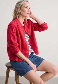 Giacca di lino rossa con colletto e bottoni frontali, indossata sopra un maglione a righe. Abbinata a pantaloni corti in denim.