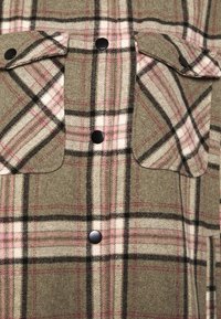 Camisa de cuadros verde hecha de una tela suave con líneas negras, blancas y rosas; cuenta con dos bolsillos y botones de presión negros.