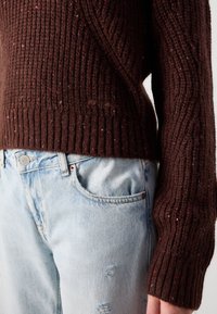 Bruine gebreide trui met geribbelde textuur, versierd met kleurrijke gespikkelde vezels, gecombineerd met versleten lichtblauwe jeans.