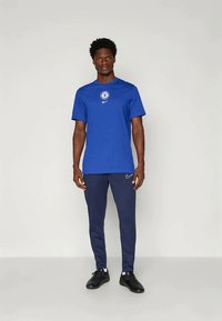 Blaues Sport-T-Shirt mit dem Chelsea FC-Logo und dem Nike-Swoosh, kombiniert mit dunkelblauen Hosen. Schwarze Sportschuhe vervollständigen das Outfit.