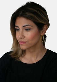Zlatna naprsna naušnica s tankim, kružnim dizajnom. Model ima tamnu kosu i nosi crnu majicu, ističući svijetlu boju naušnice.