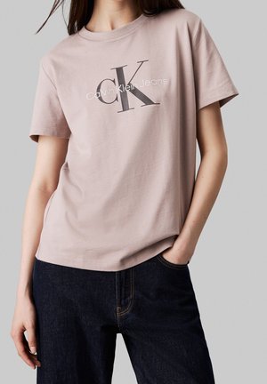 Kobieta w jasnoróżowej koszulce Calvin Klein Jeans i ciemnoniebieskich jeansach, z jedną ręką w kieszeni, na jednolitym szarym tle.