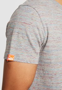 Šedá košile s krátkým rukávem a barevnými skvrnami (červená, modrá, žlutá). Na levém rukávu je malá oranžová nášivka s logem. Materiál je směs bavlny.