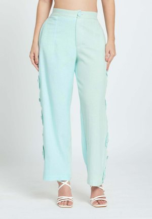 Mujer que lleva pantalones azul claro de talle alto con detalles de botones en los lados, combinados con sandalias blancas de tacón con tiras.
