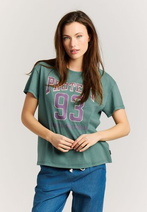 Jonge vrouw met lang bruin haar die een groen T-shirt met "Protec 93" draagt en een blauwe broek, staand met haar handen voor zich geklemd.