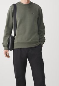 Felpa verde oliva con collo rotondo e un piccolo logo sul petto, abbinata a pantaloni neri e una borsa a tracolla nera.