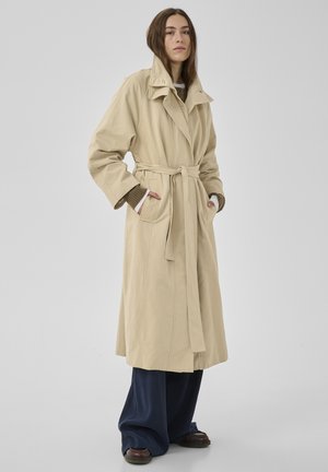 Frau trägt einen beigen Trenchcoat mit Gürtel über einem braunen Pullover und weitem, marineblauem Hosen, steht mit den Händen in den Taschen vor einem schlichten Hintergrund.