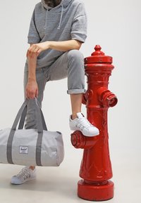 Bolso de lona gris con dos asas grises, llevado con jeans grises y zapatillas blancas, apoyado en un hidrante de incendios rojo brillante.