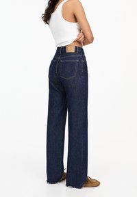Wide-leg blue jeans made of denim, featuring a small back pocket, frayed hem, and a tan leather patch on the waistband.