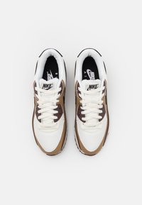 Pár bílých, hnědých a černých tenisek Nike Air Max s bílými šňůrkami, viděný z vrchu na čistě bílém pozadí.