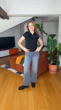 Fiatal nő fekete pólóban és szürke farmernadrágban mosolyogva áll egy modern nappaliban, nagy zöld növényekkel és fa padlóval.