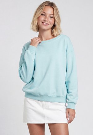 Junge Frau mit kurzen blonden Haaren lächelt, trägt einen hellblauen Pullover und einen kurzen weißen Rock, steht vor einem schlichten Hintergrund.