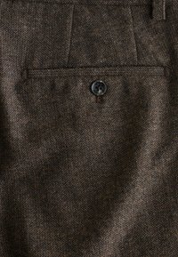 Brown tweed fabric with a herringbone pattern; features a side pocket and a black button. Texture is coarse and woven.
