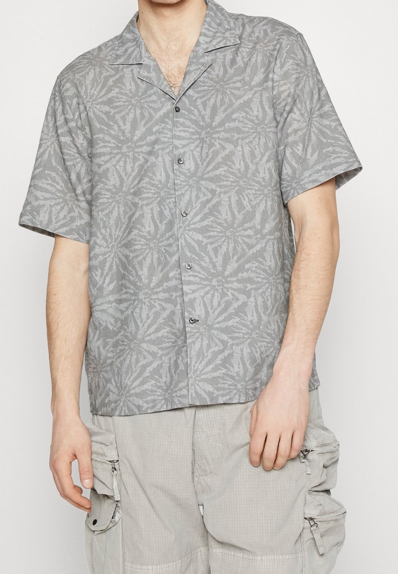 Homme portant une chemise grise à manches courtes avec un motif abstrait et un pantalon cargo gris clair avec plusieurs poches.