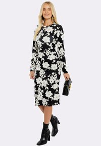 Musta kukallinen pitkähihais dressi, jossa on tiukka siluetti, valkoisilla kukkakuviolla, yhdistettynä mustiin nilkkureihin ja mustaan clutch-laukkuun.