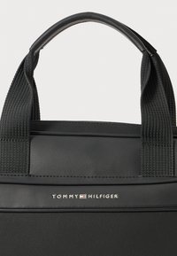 Black tote bag with a textured finish, featuring durable fabric straps and a smooth top handle. Displays "TOMMY HILFIGER" logo in silver.