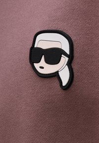 Parche de goma con un rostro estilizado, con cabello blanco y gafas de sol negras, adherido a un fondo de tela texturizada color malva.