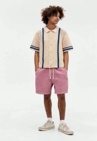 Μπεζ πλεκτό μπλουζάκι κοντομάνικο με μπλε λεπτομέρειες, με κουμπιά στο μπροστινό μέρος, συνδυασμένο με ροζ σορτσάκια με κορδόνι, και ανοιχτόχρωμα αθλητικά παπούτσια.