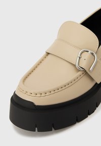 Mocassino in pelle beige con fibbia, caratterizzato da dettagli di cucitura e una suola piatta nera spessa con una finitura testurizzata.