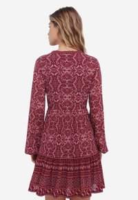 Rode jurk met patroon, lange mouwen, een getailleerde bodice en een flonkerende ruffle zoom. De stof heeft ingewikkelde bloem- en spiraalontwerpen.